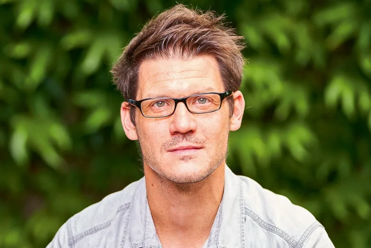 Porträtaufnahme: Ein Mann mittleren Alters mit kurzen, dunkelblonden Haaren und rechteckiger Brille schaut mit wachem Blick zur Kamera.