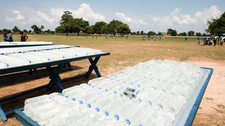 Im Vordergrund lagern abgefüllte Wasserflaschen auf Tischen, während im Hintergrund Schulkinder spielen.