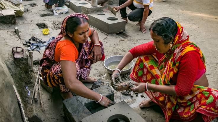 Zwei indische Frauen in roten Saris bauen gemeinsam einen Lehmherd