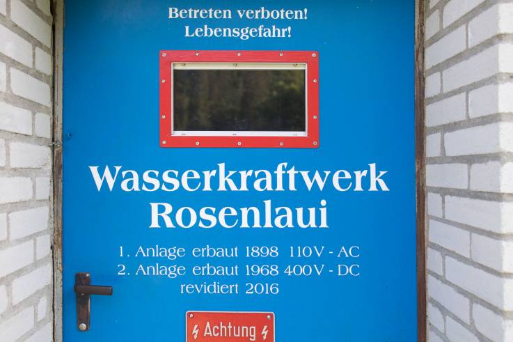 Eine robuste Stahltür mit Sichtfenster, darauf die Beschriftung «Wasserkraftwerk Rosenlaui», Warnhinweise und die Baudaten des Kraftwerks, 1898 und 1968