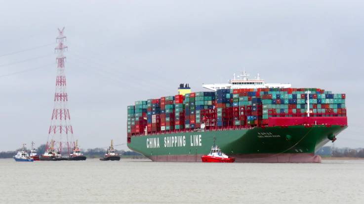 Ein riesiges , bis oben beladenes Containerschiff, umgeben von diversen Schleppern, im Hintergrund ein rot-weißer Strommast