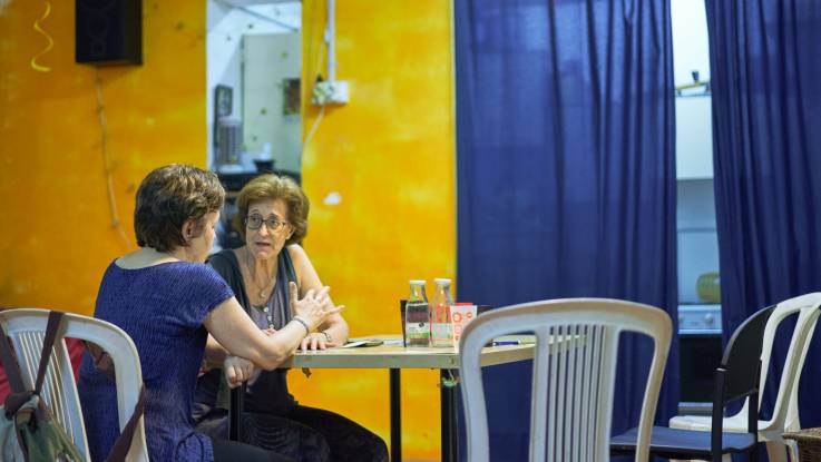 A room with yellow walls and blue curtains, two women sitting at a table chatting.