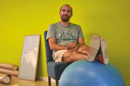 A man sits smiling on a chair, his legs resting on a blue rubber ball.