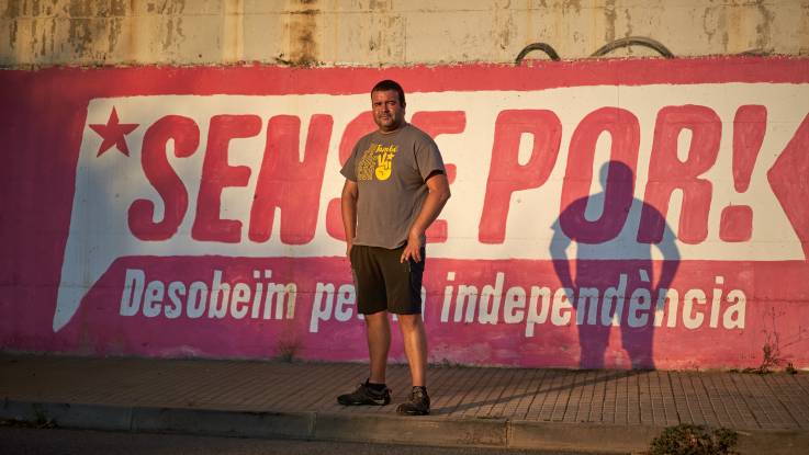 A man in a t-shirt stands in front of a wall with the slogan Sense Por! is painted on.