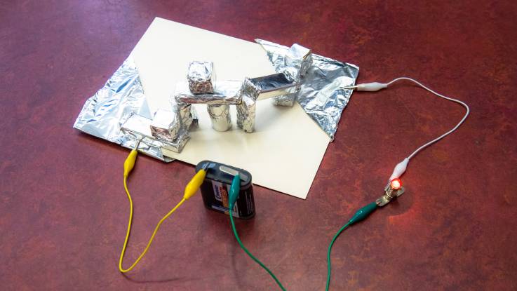 Das Experiment ist aufgebaut: Auf einer Pappe eine Konstruktion mit in Alufolien eingewickelten Bauklötzen; an den Seiten angeschlossene mit Kabel, wiederum verbunden mit einer Batterie und einer kleinen Glühbirne.