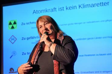 Eine Frau spricht auf einer Bühne in ein Mikrofon, im Hintergrund eine Power-Point-Folie mit der Überschrift "Atomkraft ist kein Klimaretter"