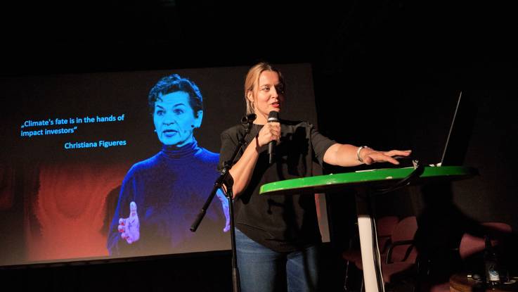 Eine junge Frau mit Mikrofon referiert auf einer größeren Bühne. Im Hintergrund eine Projektion.