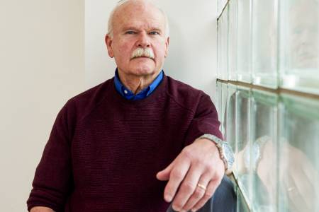 Portrait eines älteren Mannes mit Schnauzbart, der ernst in die Kamera schaut; rechts von ihm eine Wand aus Glasbausteinen.