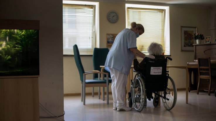 In einem abgedunkelten Raum sitzt eine alte Dame im Rollstuhl am Tisch, eine Pflegerin neben ihr.