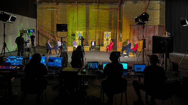 A television studio with a number of cameras and monitors has been set up in an industrial building. A group of people are sat on the stage in a semicircle having a panel discussion.