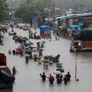 In einer überfluteten Straße laufen Menschen mit Regenschirmen durch das hüfthoch stehende Wasser, Lastwagen stehen quer. 