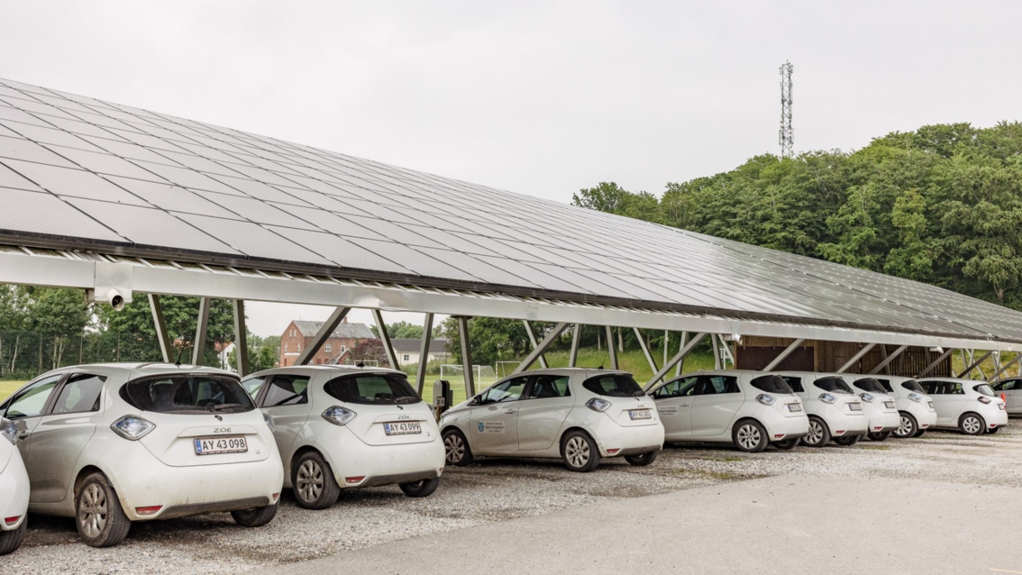 White electric cars of the same model park under a pitched roof on which PV modules are installed.