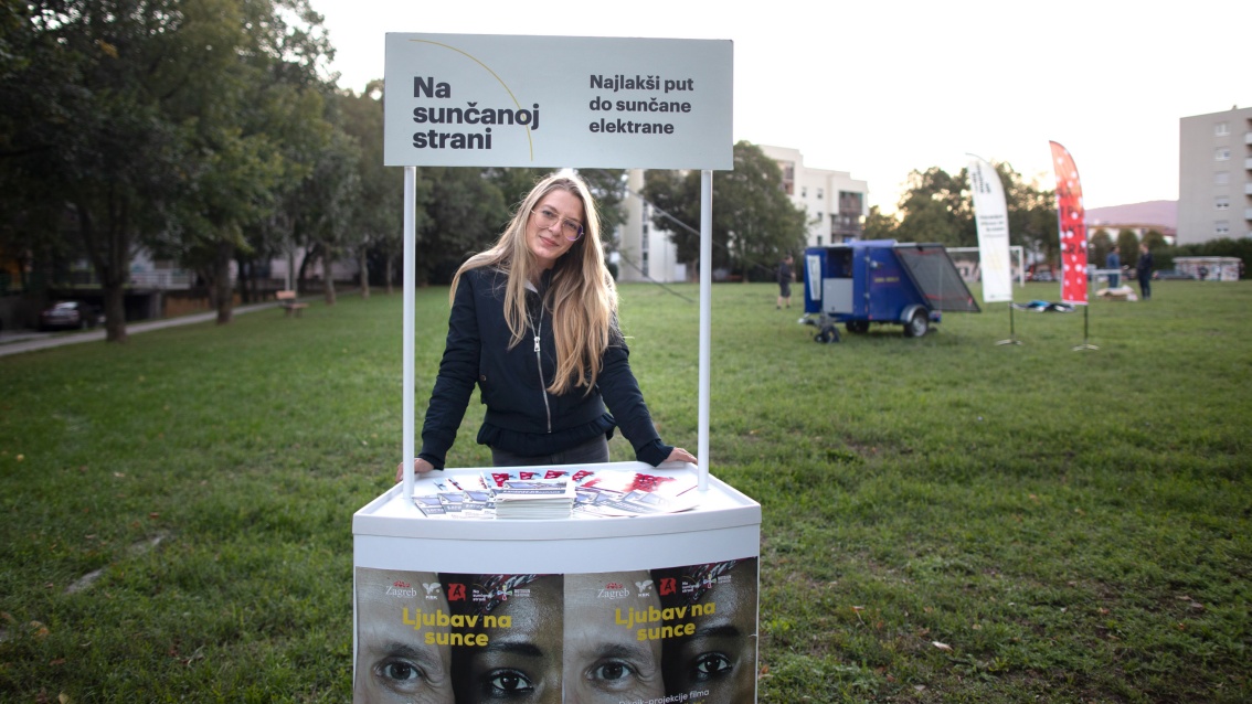 A blonde woman stands smiling and with her head tilted at an information stand, the screen is set up behind her.