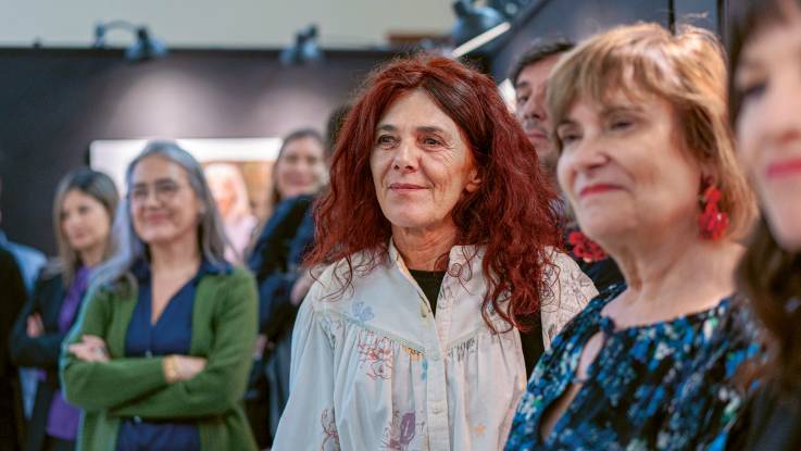 Die Frau mit den roten Haaren steht mit anderen Personen in einer Galerie und hat ein sanftes Lächeln im Gesicht.