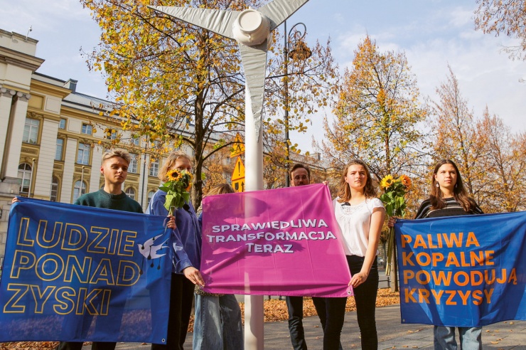 Eine Gruppe junger Menschen hält drei Banner mit verschiedenen Aufschriften, zwischen ihnen steht eine Windrad-Attrappe. 