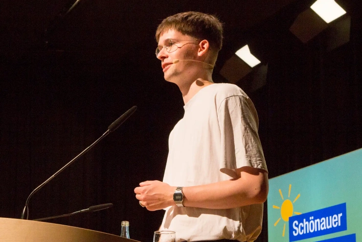 Ein junger Mann mit Brille und naturweißem T-Shirt steht an einem Rednerpult, hinter ihm eine Leuchtstele mit der Aufschrift «Schönauer Stromseminar»