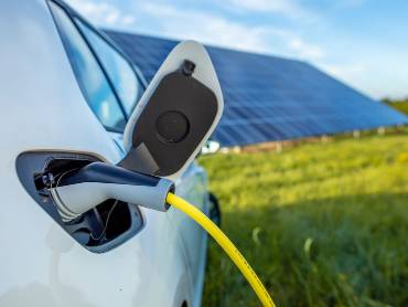 Vor einem Fld mit einer großen PV-Anlage steht ein weoßes Auto, das gerade mit Strom geladen wird.