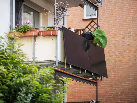 Auf dem Balkon eines Mehrfamilienhauses steht ein Mann und winkt; auf  dem Dach über ihm sind zahlreiche Photovoltaikmodule installiert.