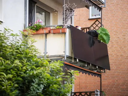 Am Balkon eines mit reichlich Pflanzen geschmückten Balkons ist eine Balkon-PV-Anlage montiert. Im Hintergrund eine Backsteinmauer.