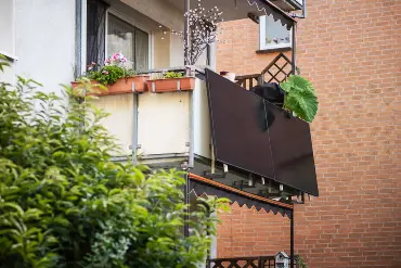 Am Balkon eines mit reichlich Pflanzen geschmückten Balkons ist eine Balkon-PV-Anlage montiert. Im Hintergrund eine Backsteinmauer.