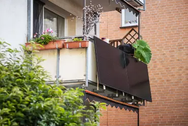 Am Balkon eines mit reichlich Pflanzen geschmückten Balkons ist eine Balkon-PV-Anlage montiert. Im Hintergrund eine Backsteinmauer.