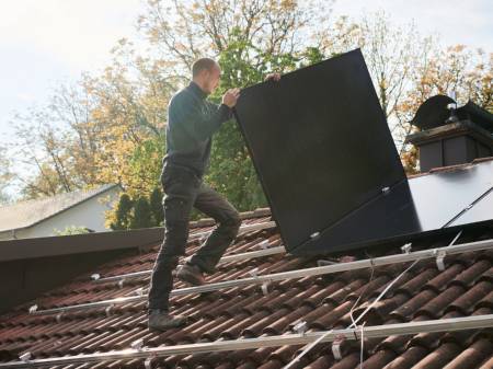 Ein Solateur auf einem Dach trägt ein PV-Modul, darauf sind Monatgeschienen, auf denen bereits viele Module installiert sind