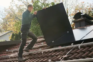 Ein Solateur auf einem Dach trägt ein PV-Modul, darauf sind Montageschienen, auf denen bereits viele Module installiert sind