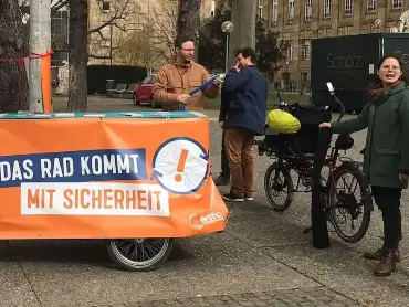 Um einen Infostand des ADFC vor einem öffentlichen Gebäude versammeln sich einige Menschen. 