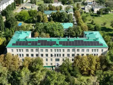 Luftaufnahme: Ein Krankenhaus, umgeben von einer Grünfläche und umrundet von mehrgeschossigen Wohngebäuden. Auf dem Dach PV-Module.