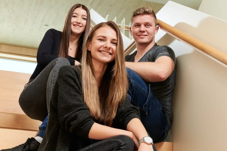 Drei junge Mitarbeitende sitzen auf einer Treppe in einem lichten Gebäude und lächeln in die Kamera.