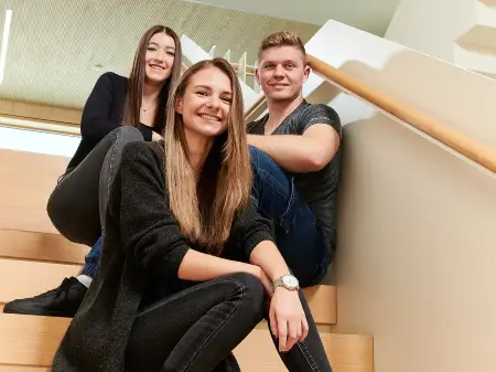 Dreii junge Mitarbeitende sitzen auf einer Treppe in einem lichten Gebäude und lächeln in die Kamera.