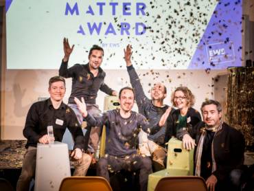 Sechs Menschen sitzen auf einer Bühne und feiern bei fliegendem Konfetti die Preisverleihung