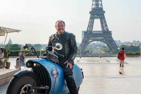 Ein E-Motorradfahrer posiert mit seiner Maschine auf einem großen Platz, im Hintergrund der Eiffelturm.