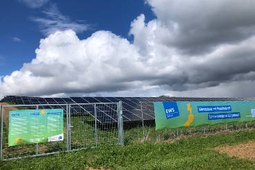 Umzäunte Solarmodule stehen auf einem Feld unter bewölktem Himmel, an den Zäunen hängen Transparente