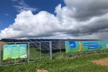 Umzäunte Solarmodule stehen auf einem Feld, an den Zäunen hängen Transparente