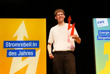 Ein lächelnder junger Mann mit Brille auf einer Bühne; in der Hand eine blitzförmige Trophäe, im Hintergrund ist auf einem Display «Stromrebell:in des Jahres» zu lesen.