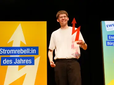 Ein lächelnder junger Mann mit Brille auf einer Bühne; in der Hand eine blitzförmige Trophäe, im Hintergrund ist auf einem Display «Stromrebell:in des Jahres» zu lesen.
