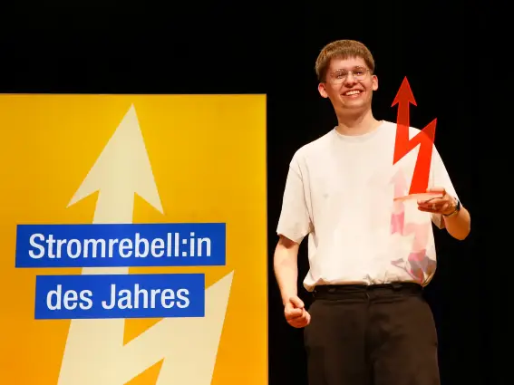 Ein lächelnder junger Mann mit Brille auf einer Bühne; in der Hand eine blitzförmige Trophäe, im Hintergrund ist auf einem Display «Stromrebell:in des Jahres» zu lesen.