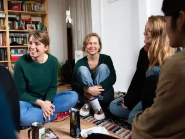 In einem Wohnzimmer mit großem Bücherregal sitzt eine Gruppe junger Menschen zwanglos auf dem Boden und unterhält sich fröhlich und angeregt.