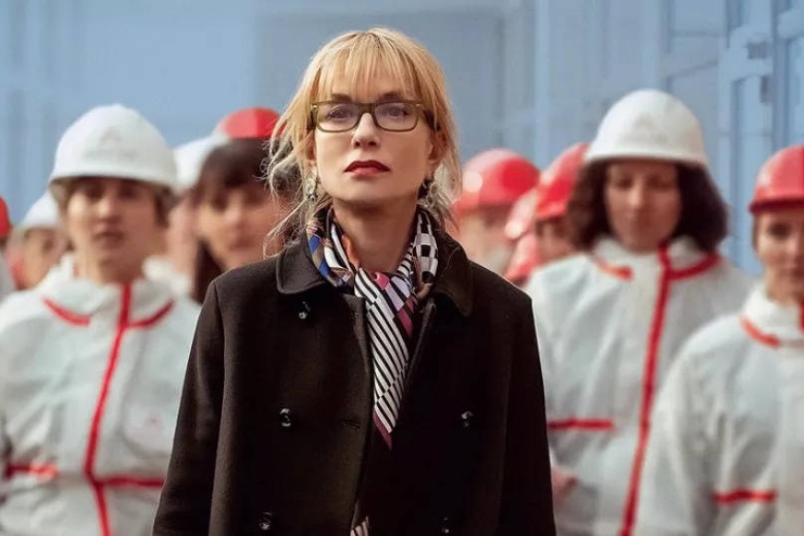 Standbild aus dem Film Die Gewerkschafterin, eine schwarz gekleidete Frau steht vor Arbeiterinnen in Sicherheitskleidung