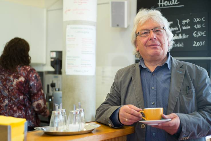 Ein lächelnder älterer Herr steht mit einer Tasse in einer  Cafeteria.