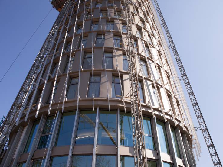 Zylindrisches Hochhaus im Bauzustand; rund um die Fassade ist eine vertikale Unterkonstruktion angebracht.