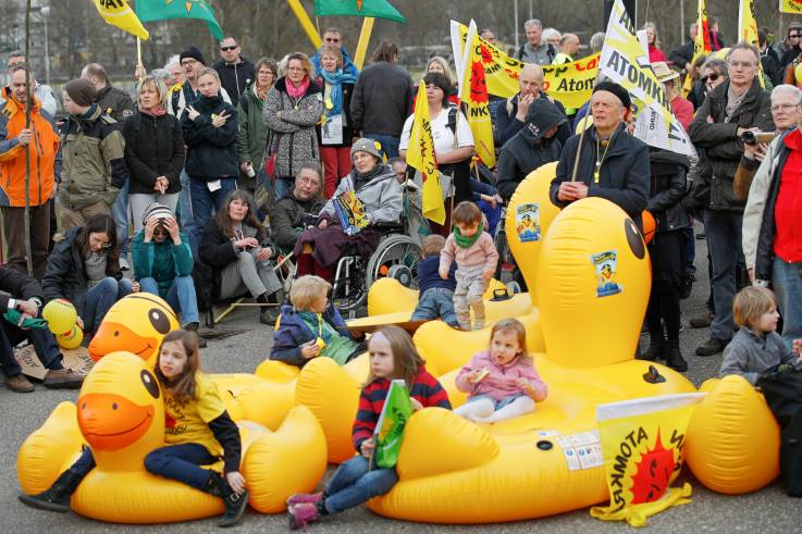 Demonstrierende mit Fahnen im Hintergrund, davor sitzen einige Kinder auf übergroßen aufgeblasenen Schwimmenten.