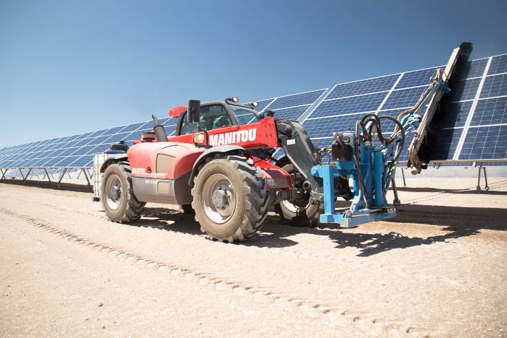 Ein Traktor fährt an den PV-Modulen entlang und reinigt mit seinem bürstenbesetzten Ausleger die schrägstehenden Moduloberflächen.