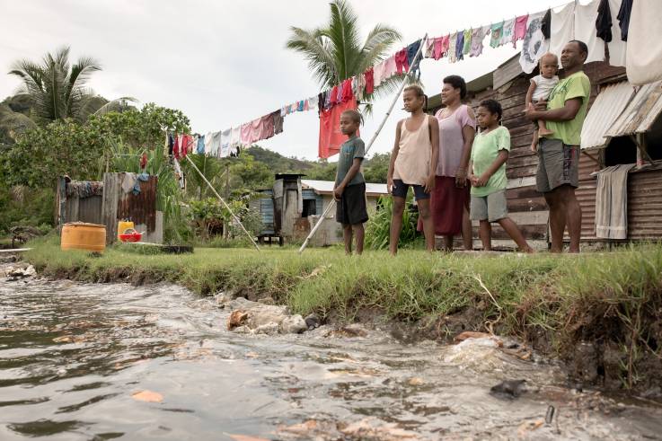 Eine Familie steht vor ihrem Haus, unmittelbar vor ihnen die durch Erosion entstandene Küstenlinie.