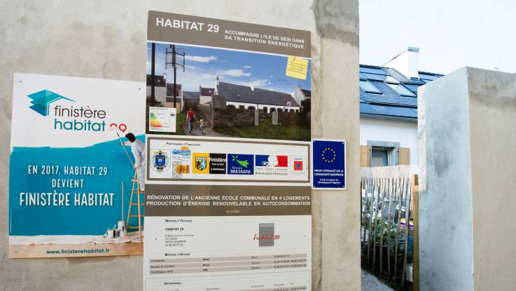 An der Mauer hängt eine Infotafel über die Photovoltaik-Anlage, die auf dem Hausdach im Hintergrund zu sehen ist.