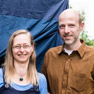 Mann mit grauem Bart und wenig Haaren und langhaarige Frau mit Brille blicken in die Kamera