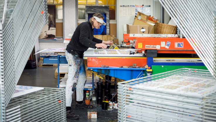 Eine Druckerei, im Vordergrund zahlreiche Trockungswagen, im Hintergrund ein Drucker mit Kopflupe