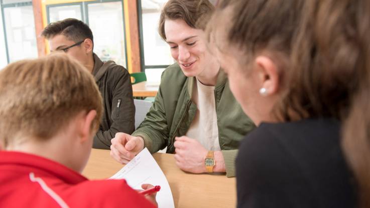 Im Vordergrund zwei jüngere Schüler, die einen Fragebogen vor sich liegen haben; im Hintergrund zwei ältere Schüler.