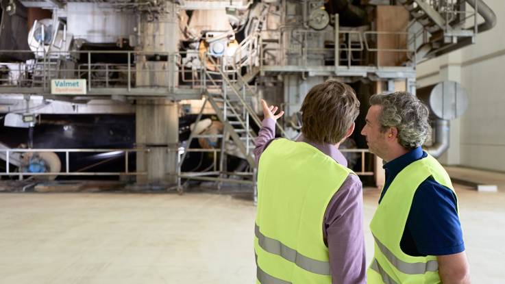 Zwei Männer in gelben Warnwesten vor einer riesigen Industrieanlage.